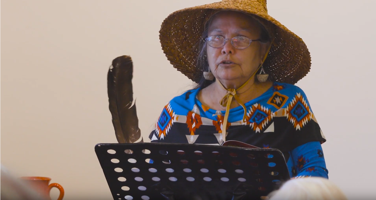 An indigenous woman in a woven hat and colourful patterned shirt reading a book passage out to a room of people